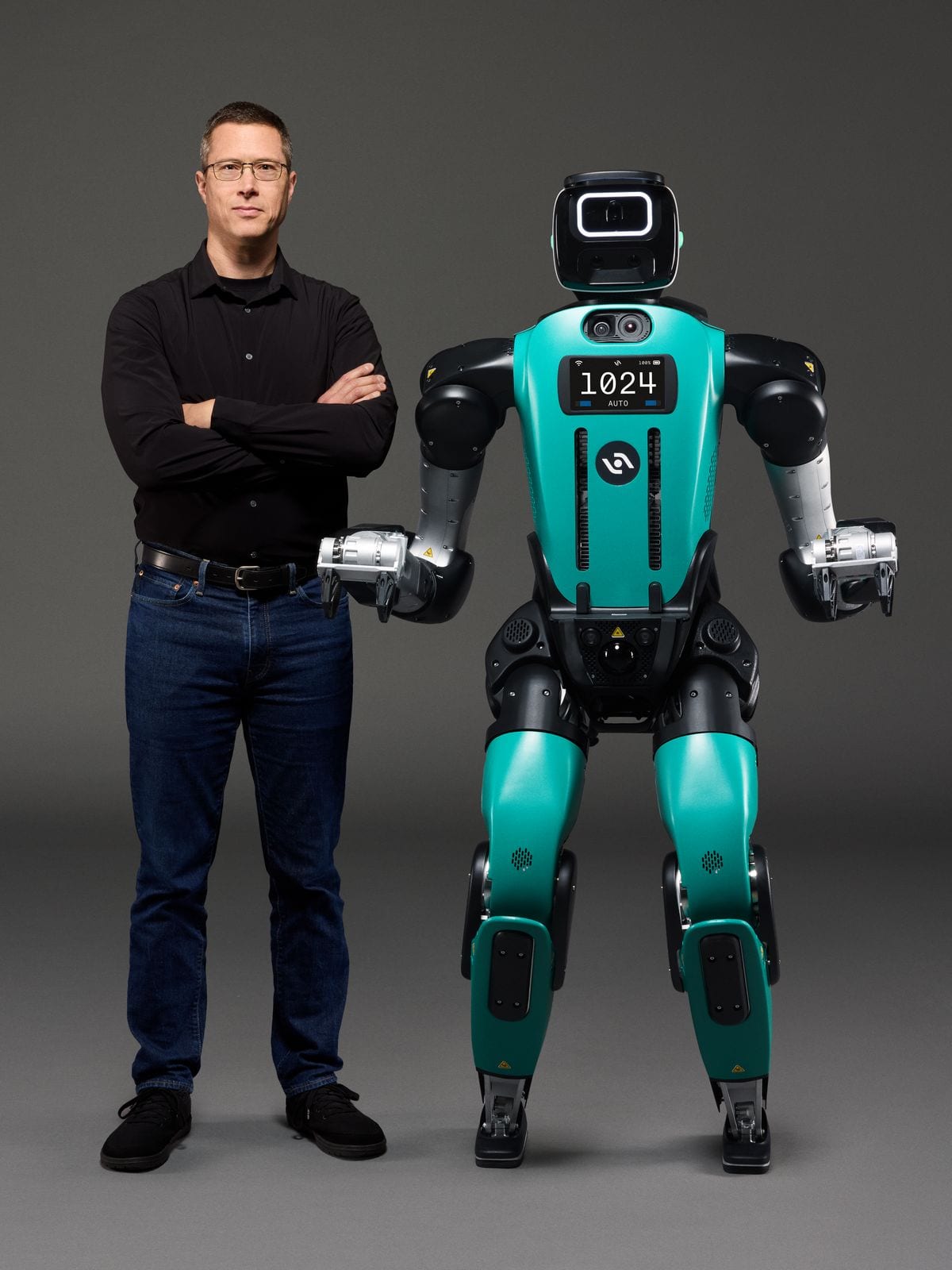 Author Jonathan Hurst, a tall slim white man wearing a black long sleeve shirt and dark jeans, stands next to his Digit 5 humanoid robot with his arms crossed. The robot is much bulkier than he is, al…