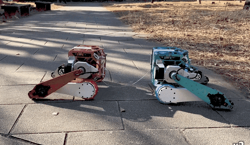 Two small robots with long limbs and bodies linked by a wire join connect to form one four-wheeled robot on a paved pathway