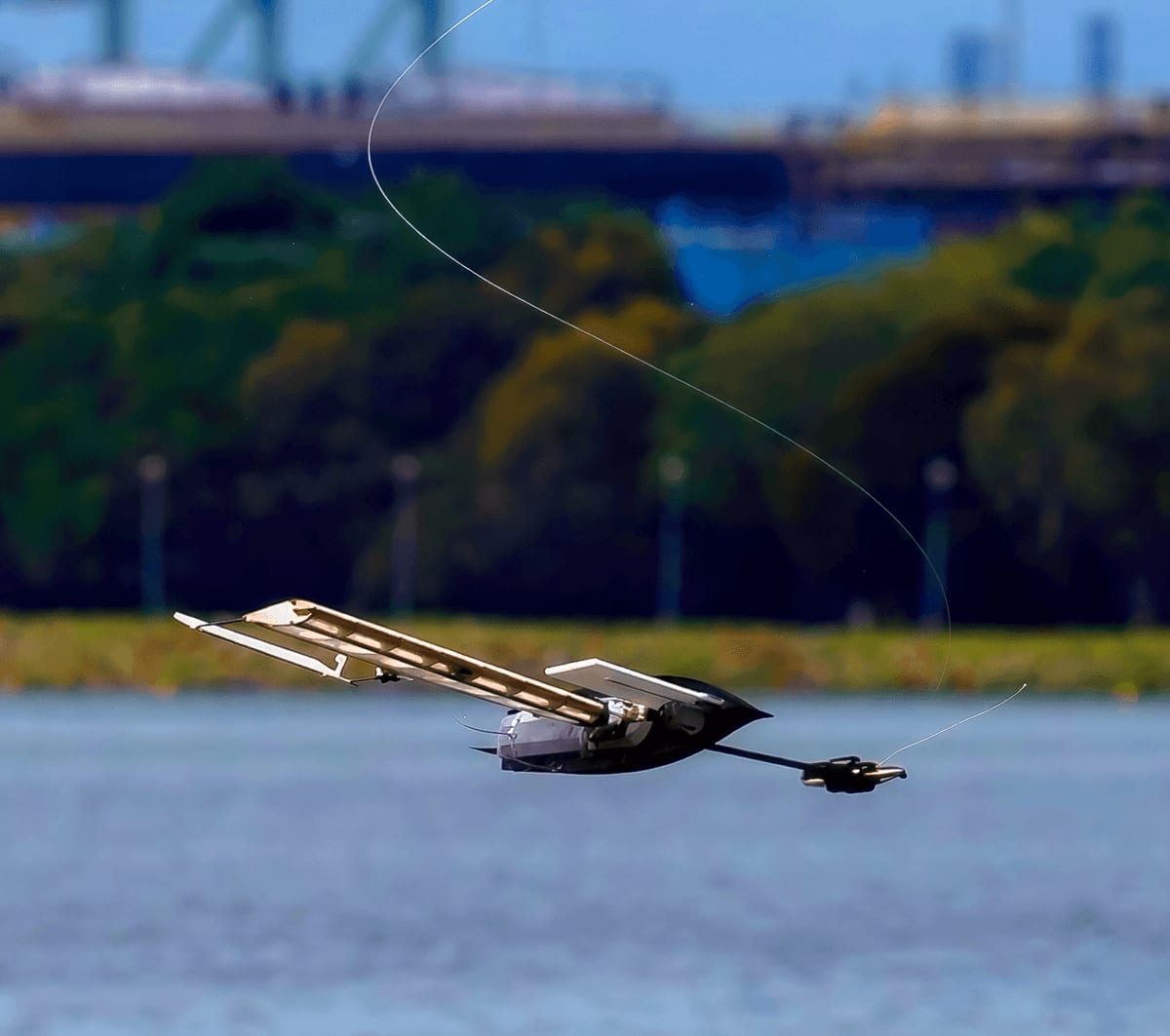 Futuristic robotic bird gliding low above water with a trailing wire.