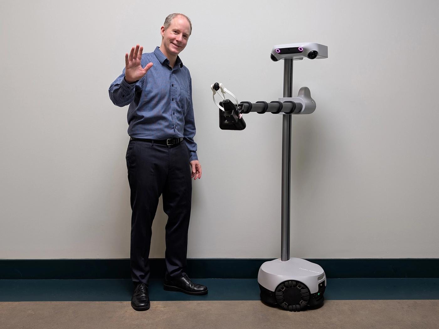 Charlie Kemp, a man in black pants and a blue shirt, smiles and waves at the camera, standing next to a Stretch 4 mobile robot consisting of a wheeled base, a tall pole, an articulated arm, and a head…