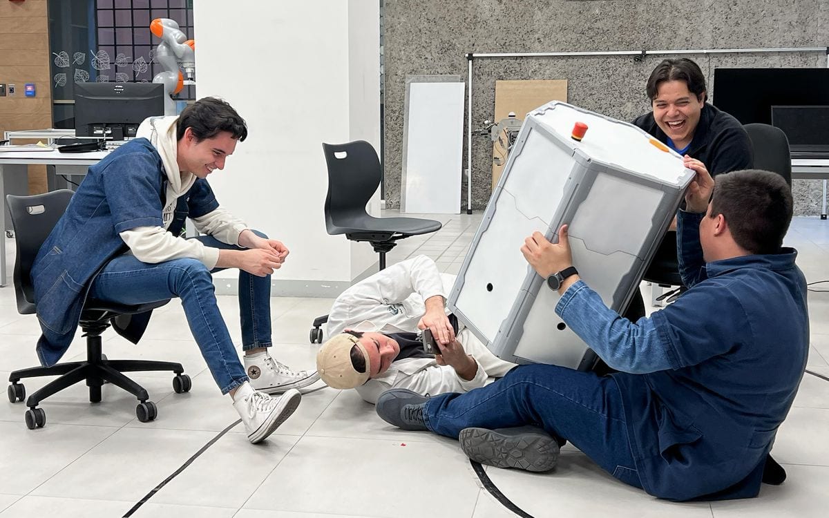 Four young adult students laugh together while one of them holds a robot shaped like a hexagonal cylinder.