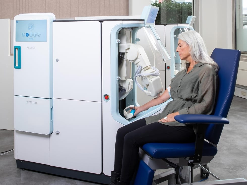 An older silver haired woman seated at automated medical device with a robotic arm drawing blood