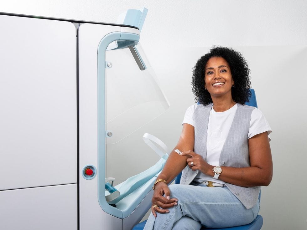 A smiling dark skinned woman holds a bandaid onto her arm while sitting next to a large machine.