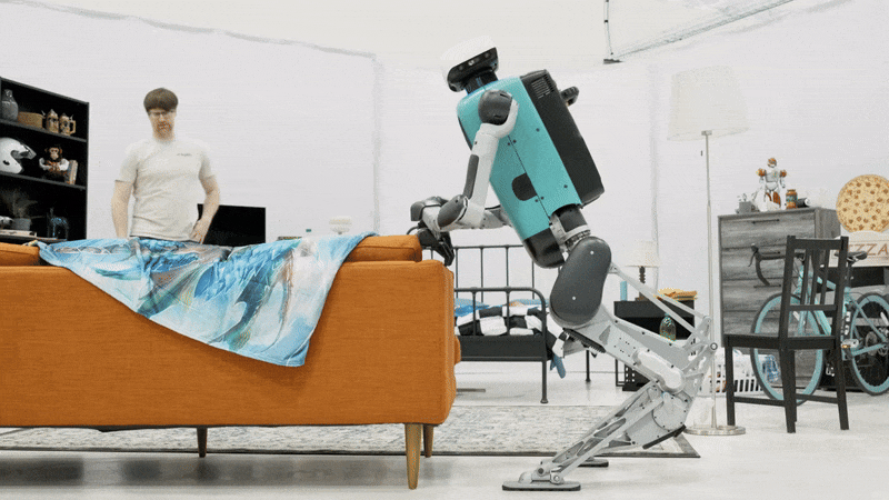 Humanoid robot tidies an orange couch while a person watches in a modern living room.