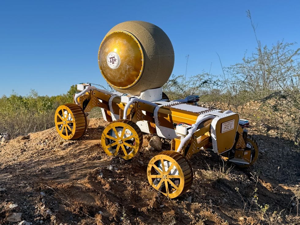 A wheeled rover carrying a robotic ball up a small hill of dirt and gravel.